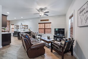A modern living room with a leather sofa, a coffee table, and a TV. There is a kitchen area with a dining table and chairs. The room has wooden flooring and a ceiling fan. - Cabin 12 @ Trails Edge 1 King 1 Queen 2bd/2ba Directly on Back 40 Trails (Bella Vista)