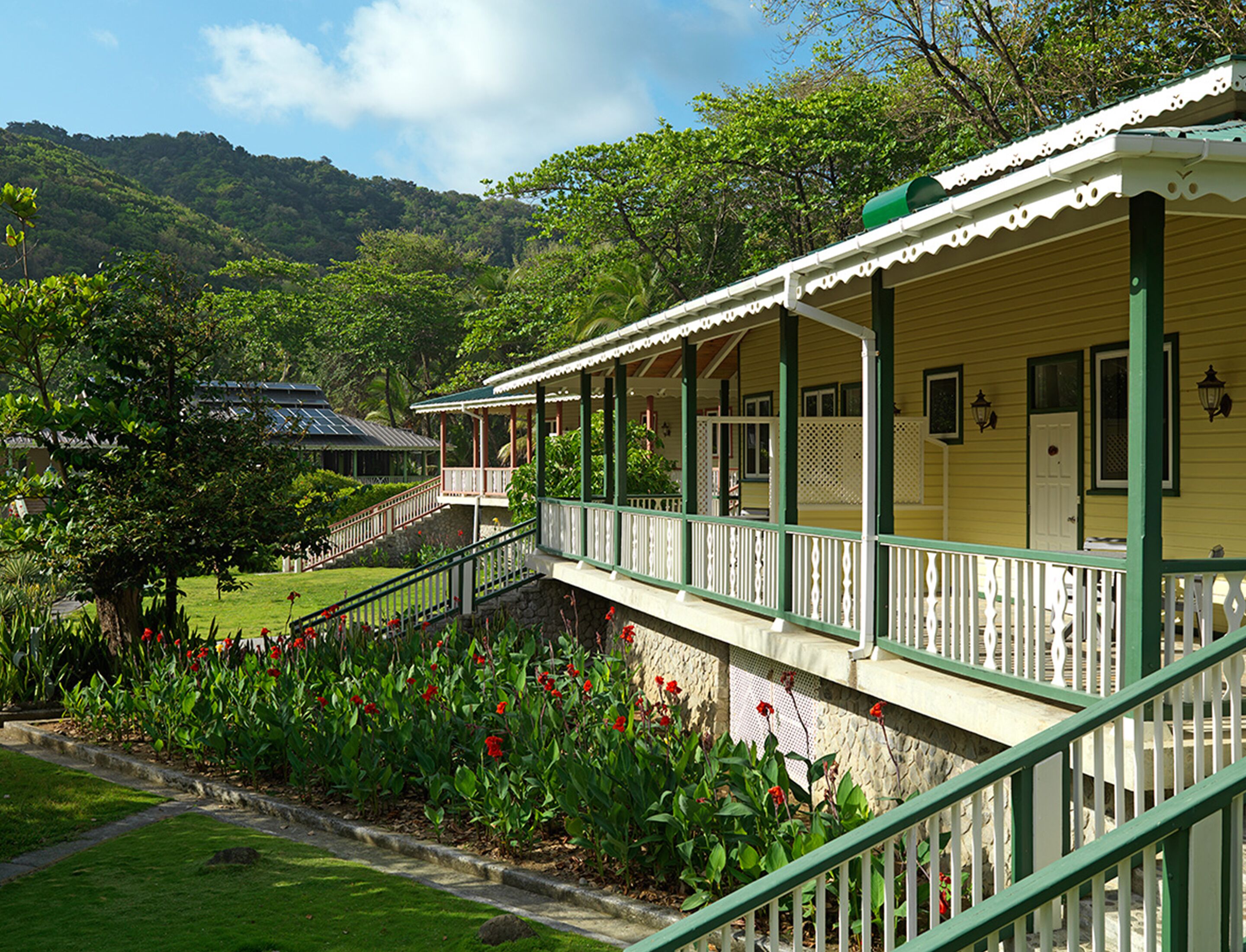 A yellow building with a green porch and white railings, surrounded by lush greenery and a stone pathway.