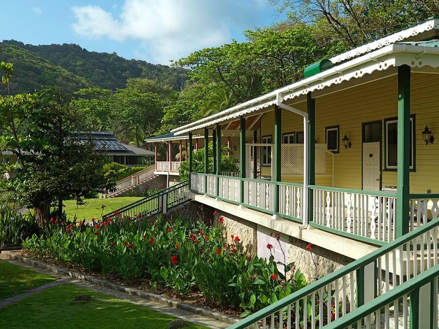 A yellow building with a green porch and white railings, surrounded by lush greenery and a stone pathway.