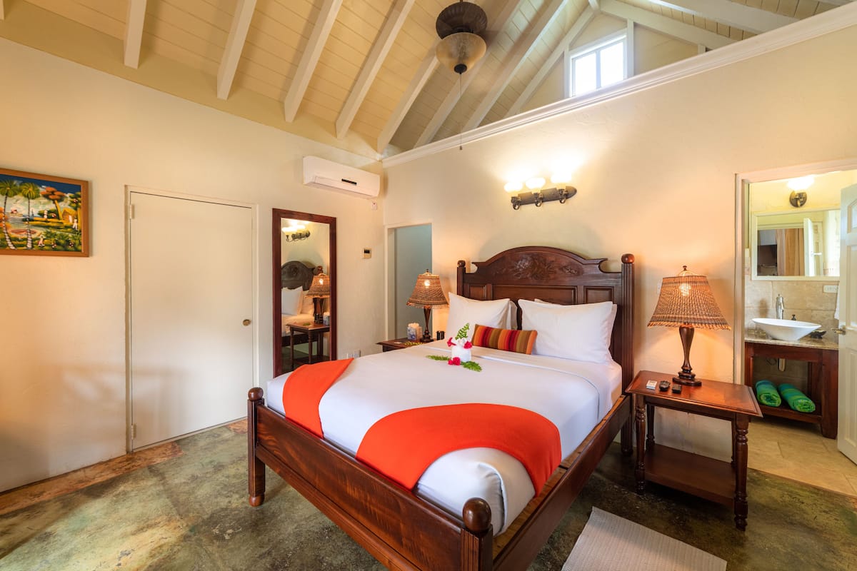 Een hotelkamer met een houten bed, een nachtkastje met een lamp, een badkamer met een wastafel en spiegel, en een schilderij aan de muur.