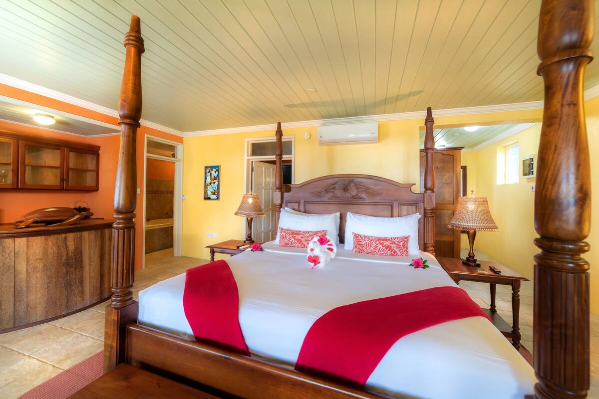 Een hemelbed met een houten hoofdeinde, een rode en witte spreideken en twee bedlampjes.