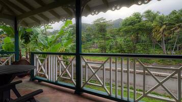 A covered outdoor area with a view of a road and lush greenery, including banana plants and palm trees.