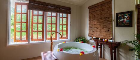 A large bathtub with a wooden chair and a small table, large windows with wooden frames, and a painting on the wall.