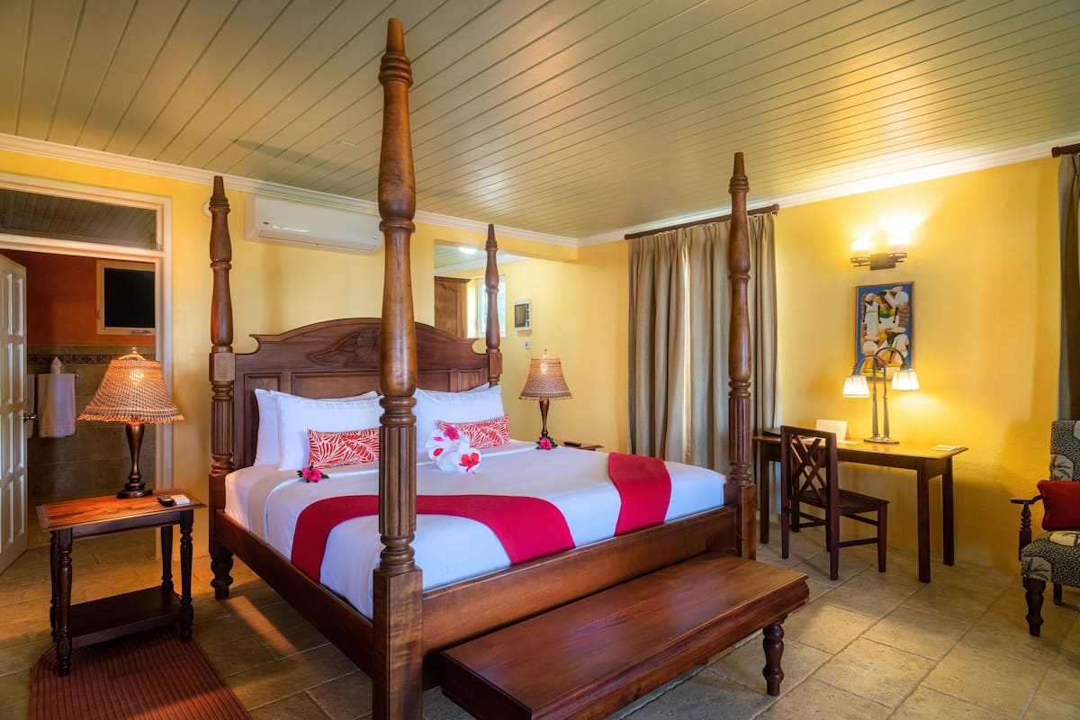 Een hemelbed met houten hoofdeinde, een bank aan het voeteneinde, een nachtkastje met een lamp en een stoel met kussen.