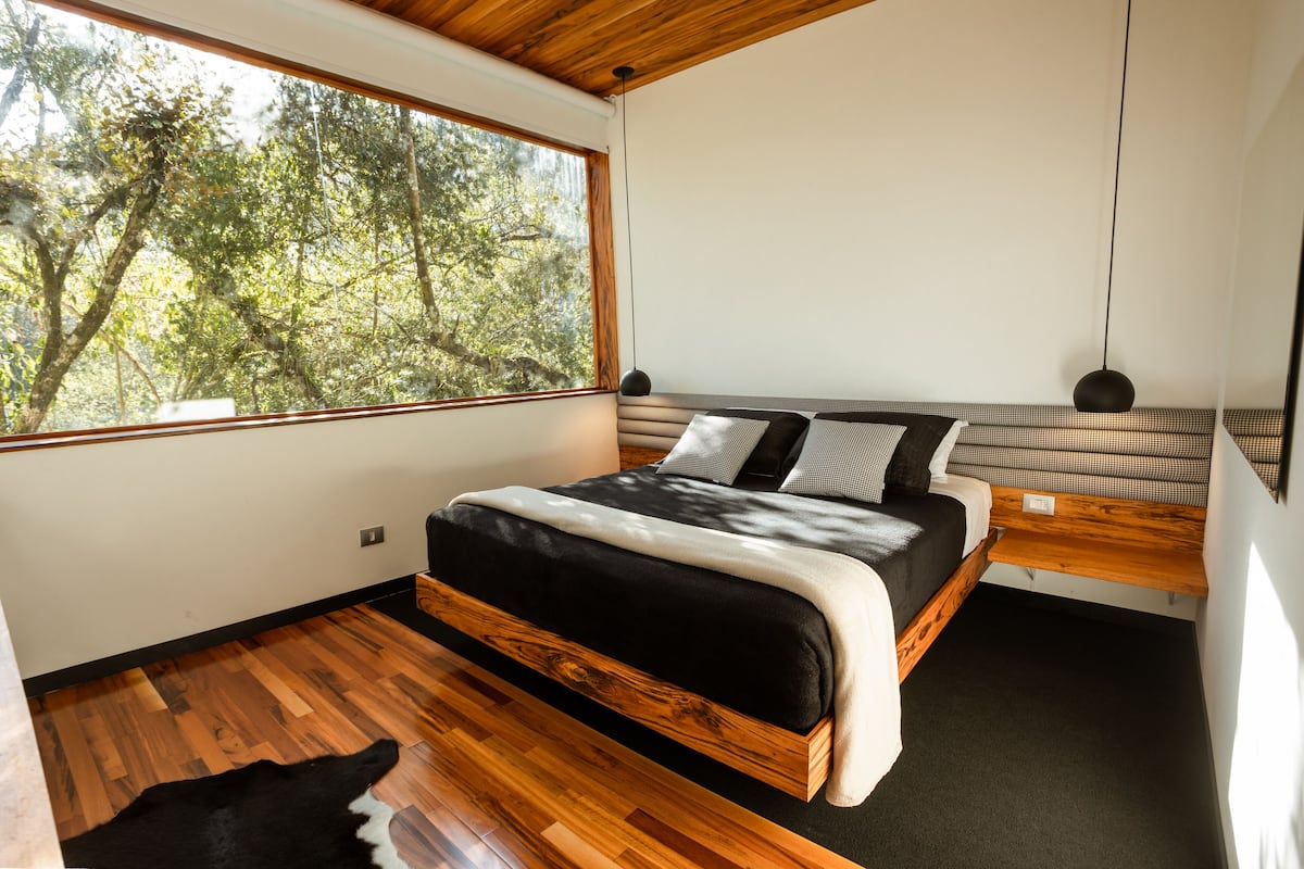 Ein modernes Schlafzimmer mit großem Fenster, einem hölzernen Bett, einer Bank mit gemusterten Kissen und einem Schreibtisch.