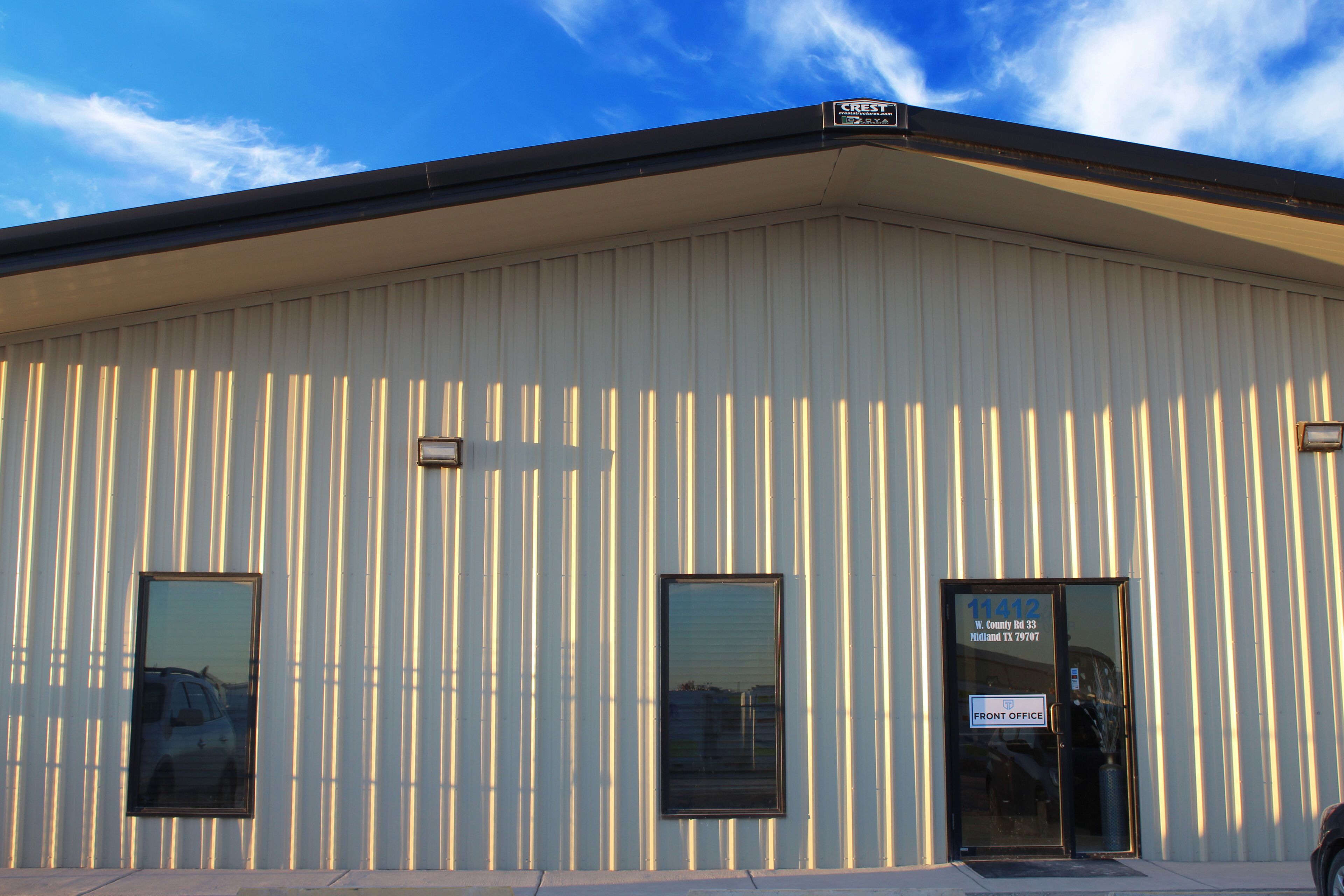 A building with vertical siding, a black door with a sign reading "11412 W. County Rd 33 Midland TX 79707 FRONT OFFICE", and two windows.