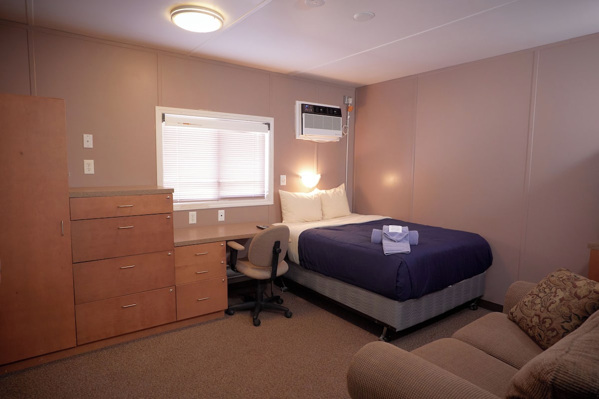 A bedroom with a bed, desk, chair, wardrobe, and an air conditioning unit.