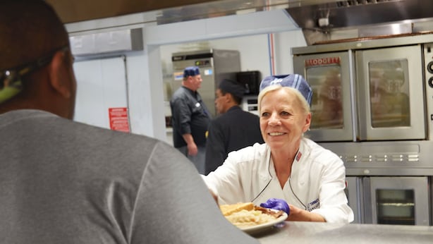 A person in a white uniform serving food in a commercial kitchen with stainless steel appliances and a visible oven. - Target Hospitality-Williams County Lodge (Williston)