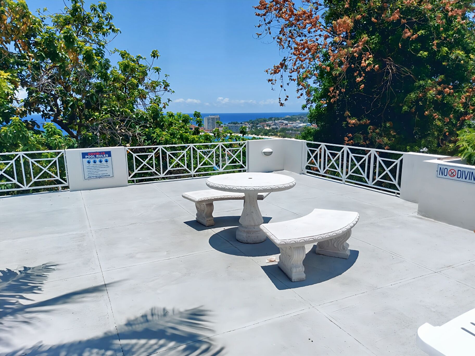 A patio with a round table and two benches, surrounded by a white railing and trees.