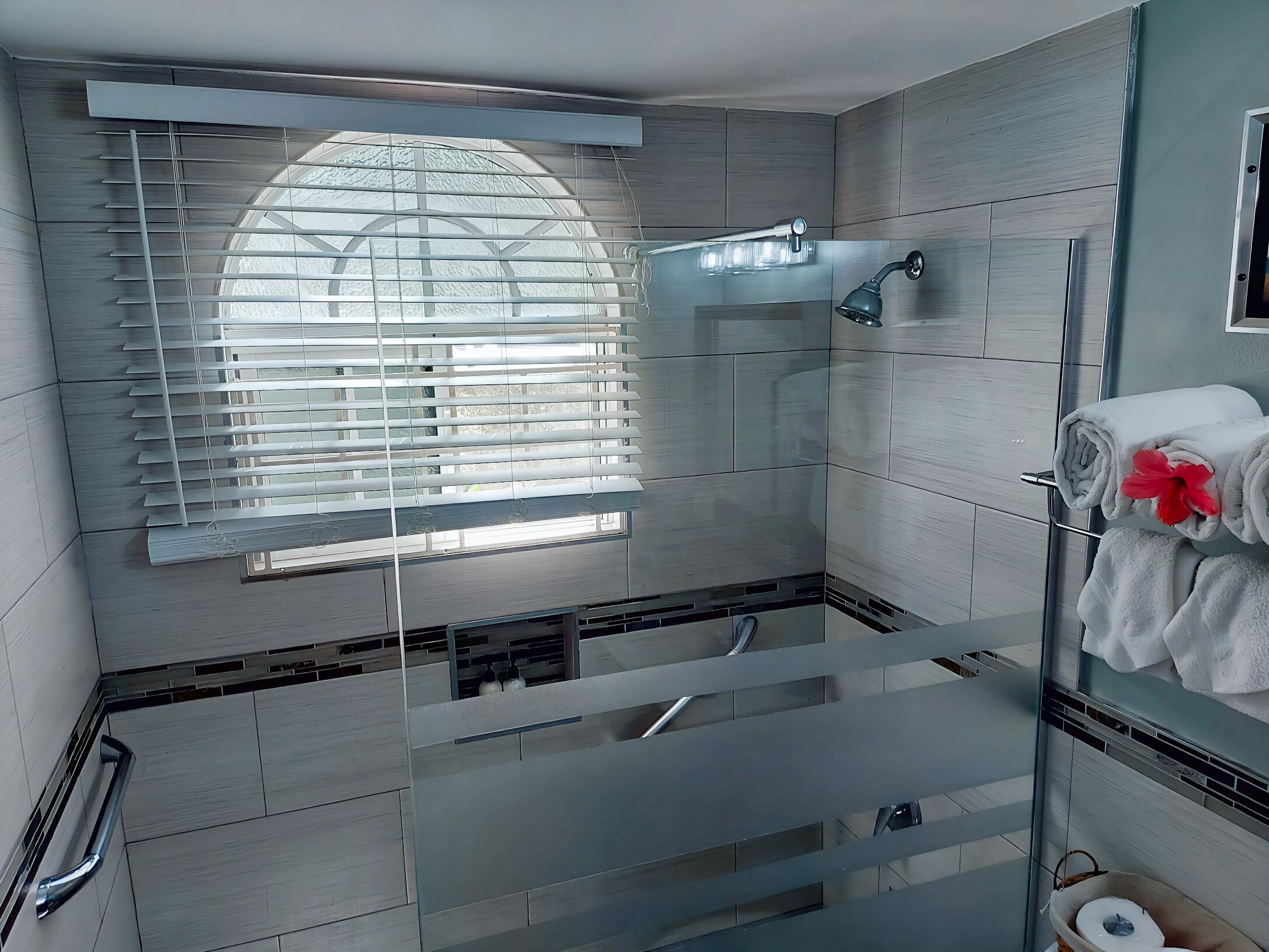 A modern bathroom with a glass shower enclosure, a built-in window with blinds, and a towel rack with towels.