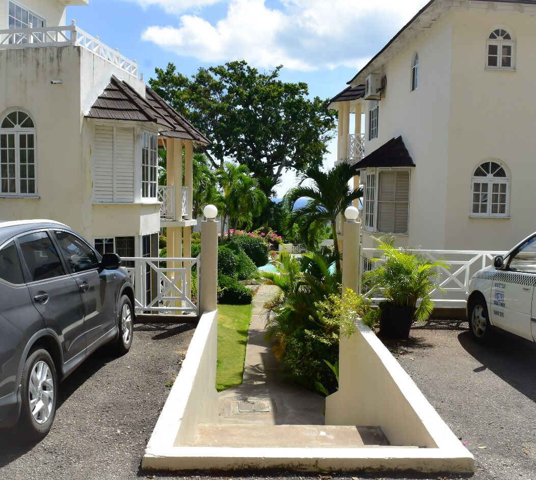 A gated entrance with a pathway leading to a building, surrounded by parked cars and lush greenery.