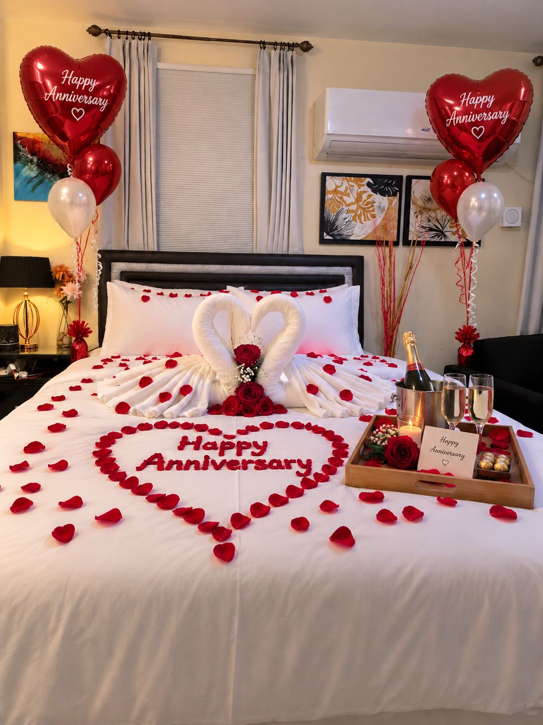 A hotel room decorated for an anniversary with a bedspread featuring a heart-shaped arrangement of rose petals and a 'Happy Anniversary' message, a bouquet of red roses, heart-shaped towels, and two heart-shaped balloons.