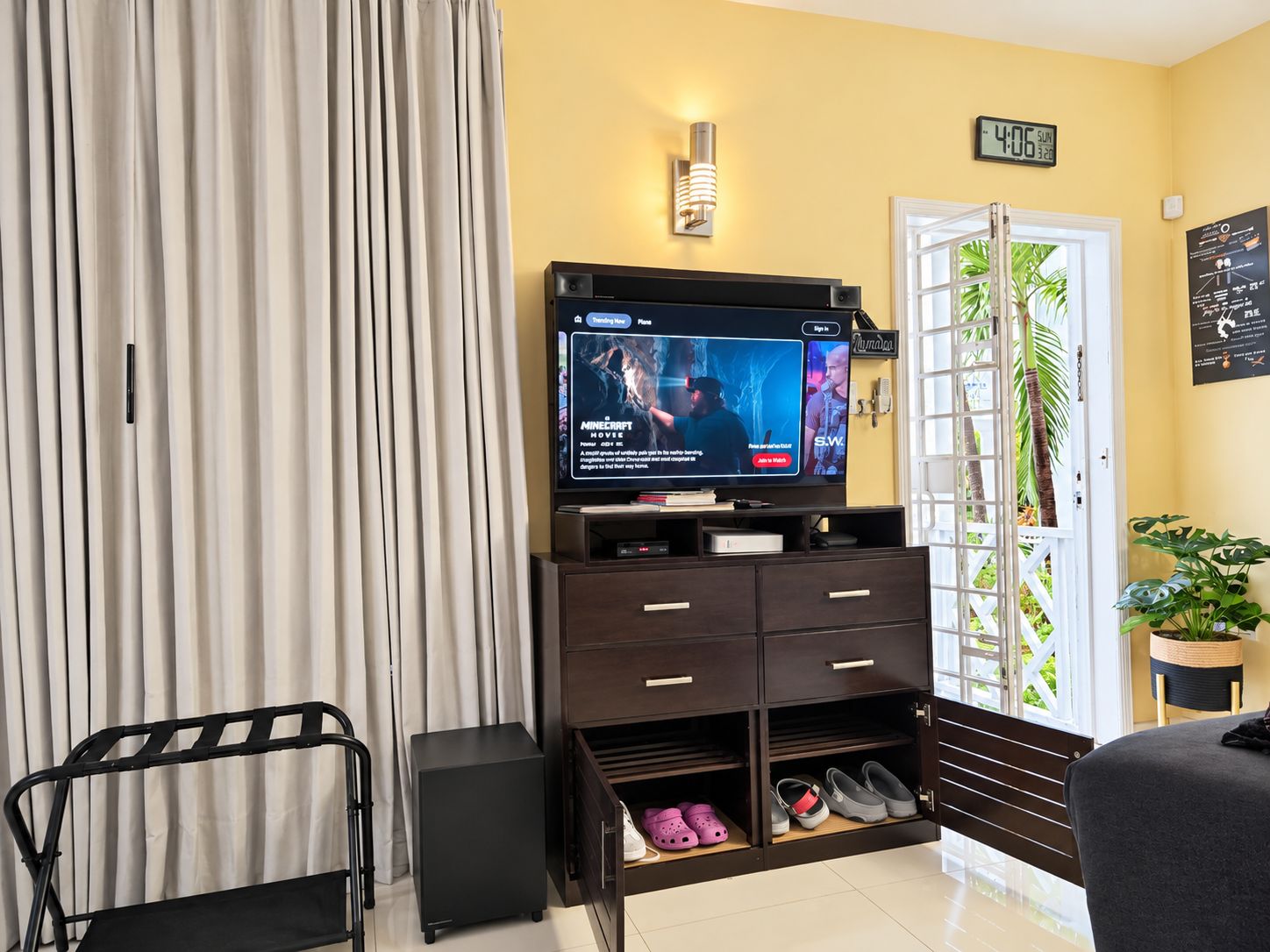 A living room with a flat-screen TV displaying a movie, a dark wooden cabinet with drawers, a black speaker, and a door with a view of greenery outside.