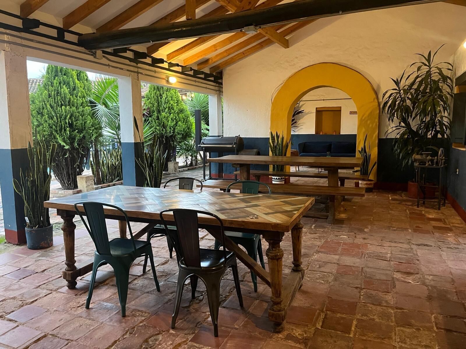 Une terrasse extérieure avec une table et des chaises en bois, entourée de plantes en pot et d’une arche jaune.