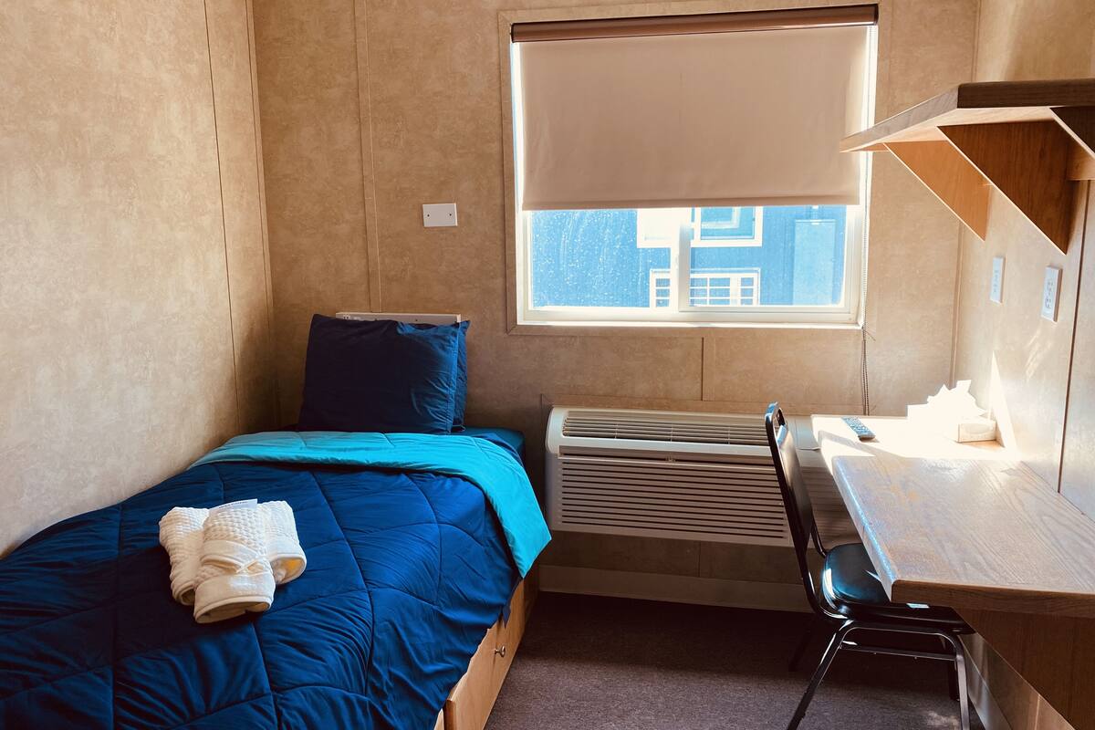 A bedroom with a bed, a desk, a chair, a window, and an air conditioning unit.