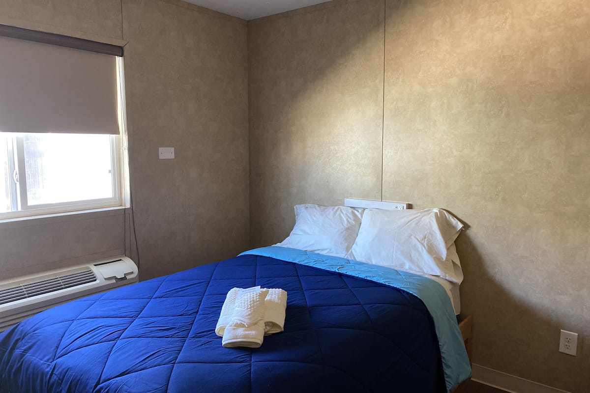 Habitación de hotel con una cama grande, una ventana con persianas, aire acondicionado montado en la pared y un enchufe.