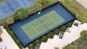 Luftaufnahme eines Tennisplatzes mit umliegender Grünfläche und einem Parkplatz.