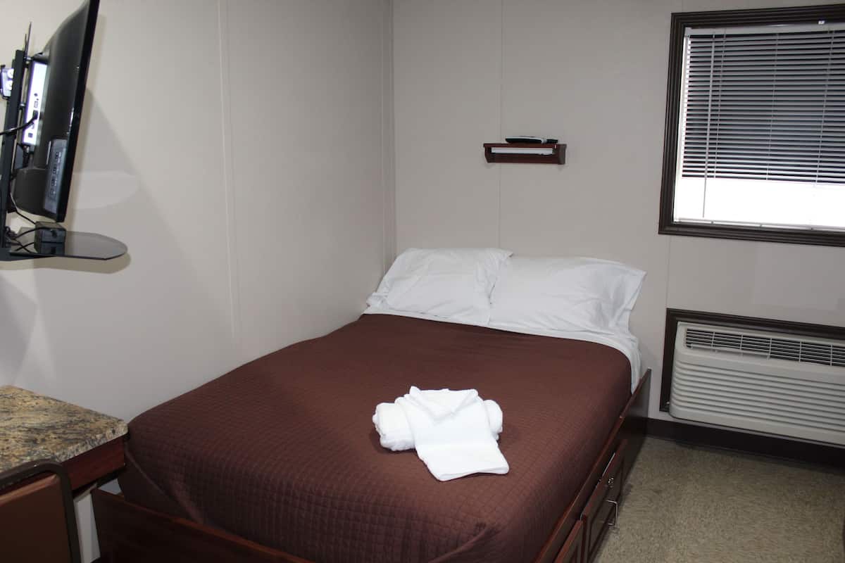 Una cama bien hecha con una colcha marrón y almohadas blancas, una mesita de noche con un televisor y una ventana con persianas.