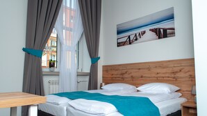 A hotel room with a wooden headboard, a bed with white linens and a teal throw, a wooden nightstand, and a window with curtains. - Villa Centro (Oswiecim)