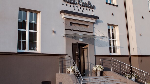 A building with a sign reading "Villa Corso Apartments." It features a set of stairs leading to the entrance, outdoor seating, and a clear sky. - Villa Centro (Oswiecim)