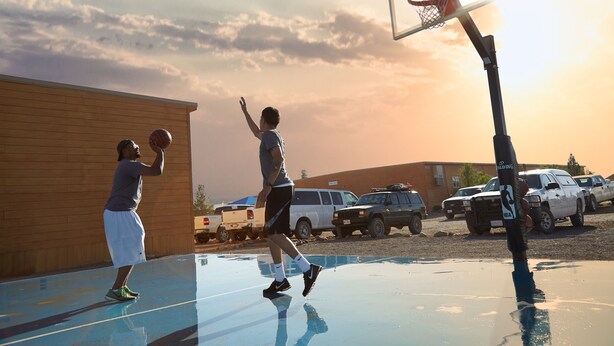 Un terrain de basketball en plein air, avec un bâtiment en bois en arrière-plan et des véhicules garés.