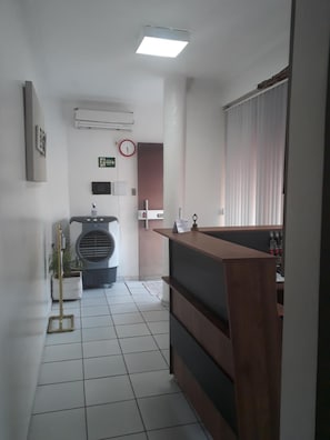 A reception desk with a clock and emergency exit sign on the wall, a radiator, and a potted plant in a hallway. - Pousada Executiva (Teresina)