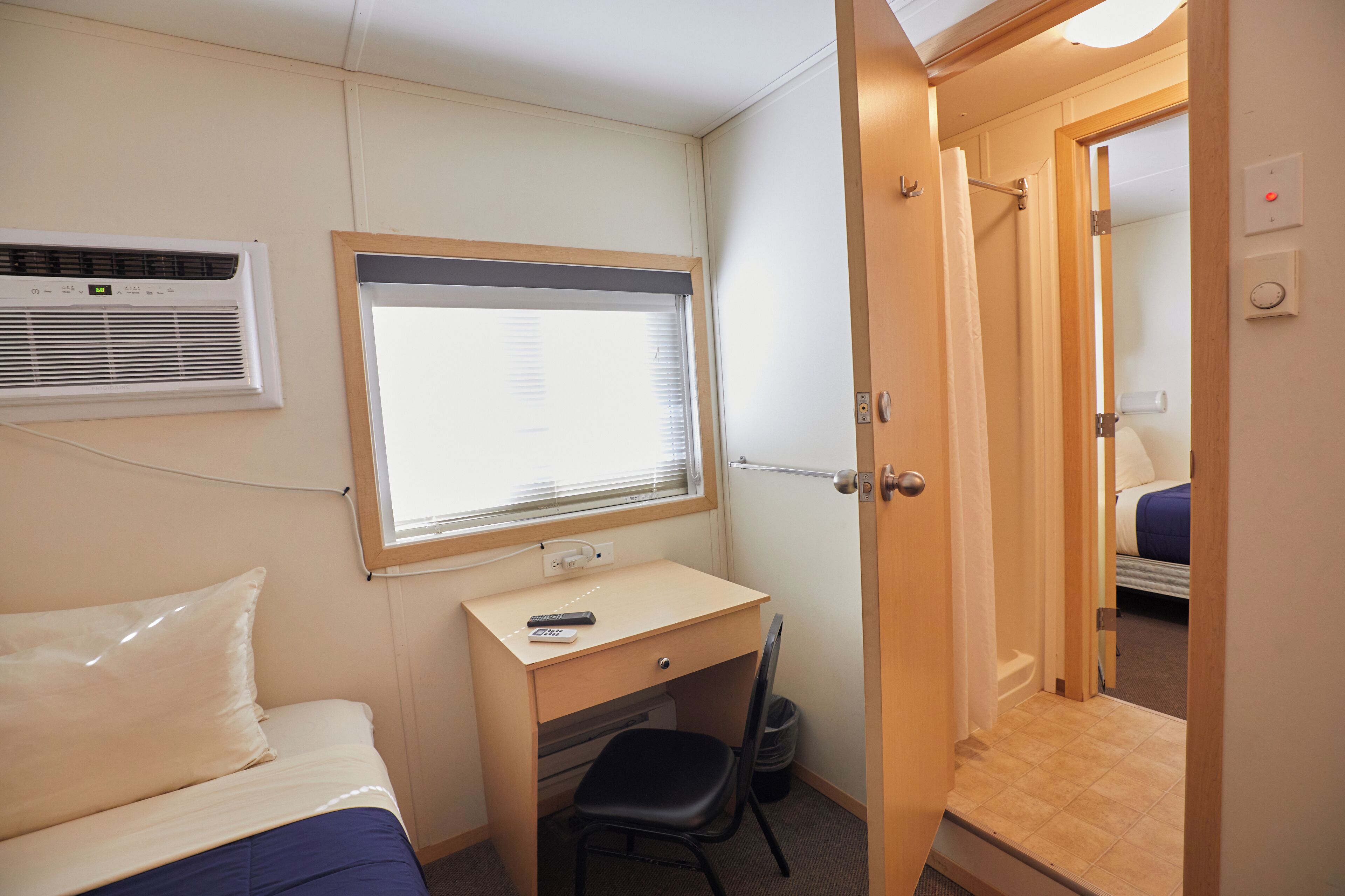 Habitación de hotel compacta con cama, escritorio y silla, ventana con persianas, unidad de aire acondicionado y puerta que da a otra habitación.