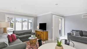Una sala moderna con un sofá gris, un sillón gris más pequeño, un mueble de madera con un televisor, y un comedor con mesa y sillas.