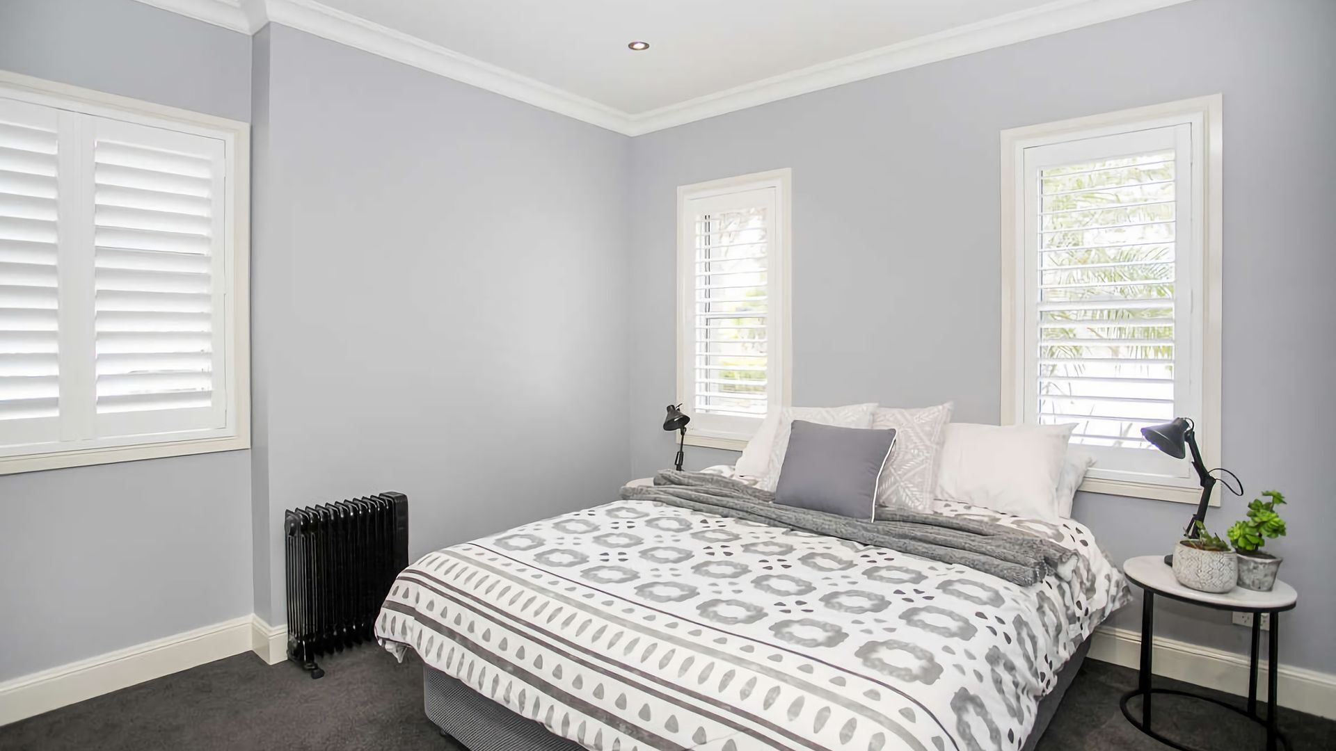 A bedroom with a bed, two bedside lamps, a radiator, and a small table with a plant.