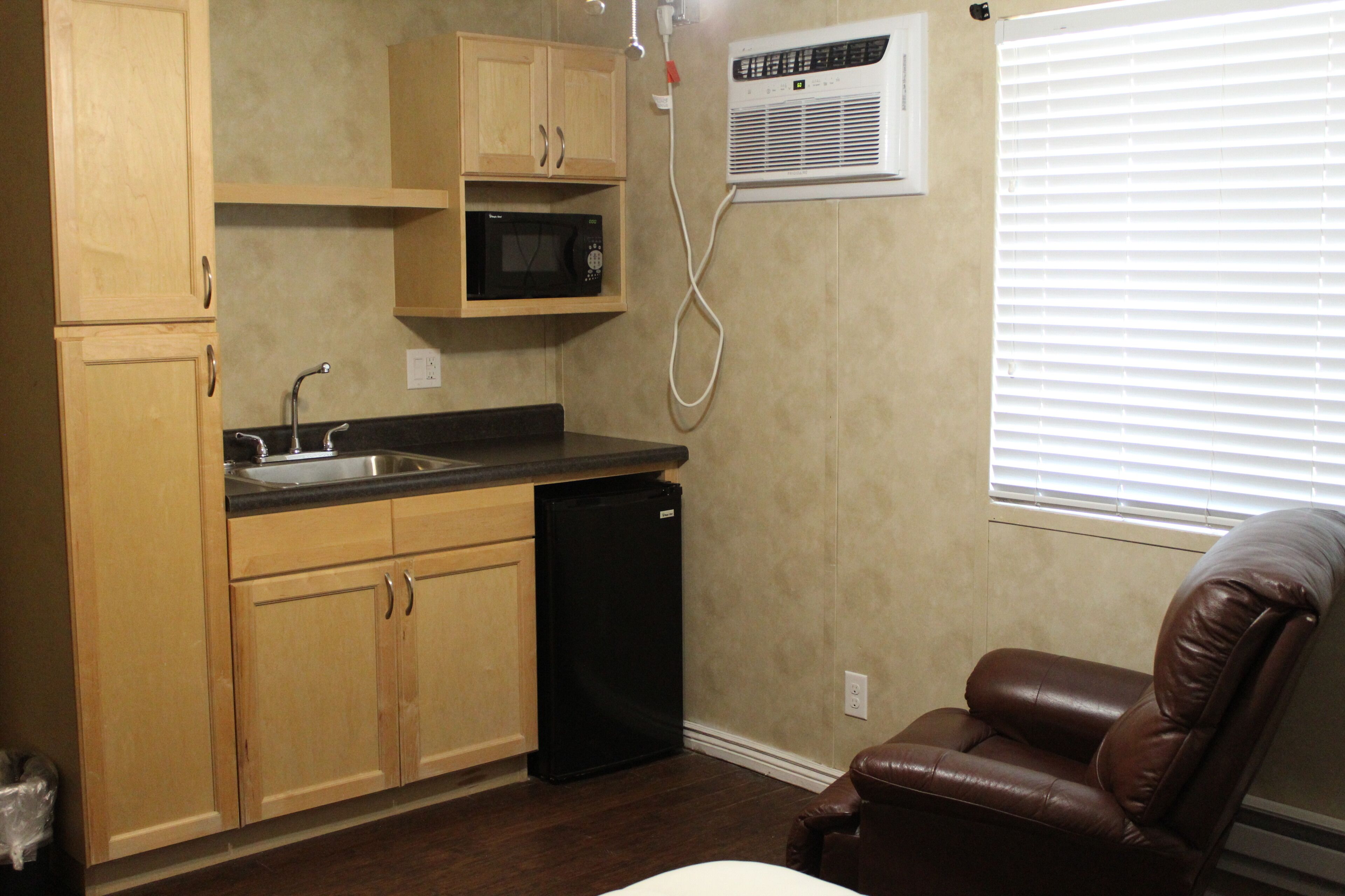 A compact kitchen with wooden cabinets, a black refrigerator, and a microwave. There is a brown leather chair and a window with blinds.