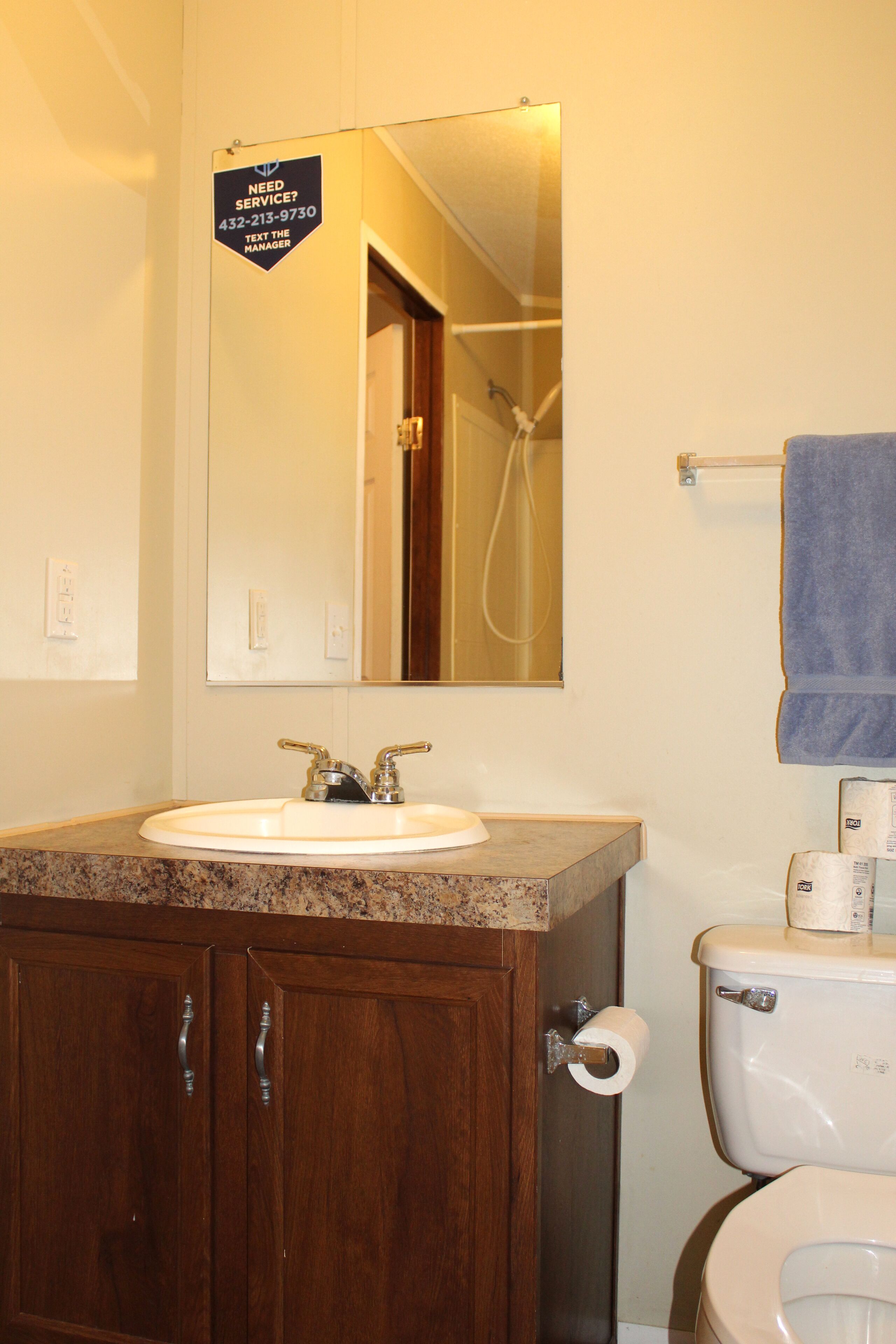 A bathroom with a sink, mirror, and toilet. There is a sign above the mirror that says "Need Service? 432-213-9730 Text the Manager."