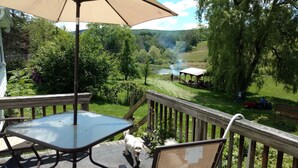 A wooden deck with a glass table and chairs, overlooking a grassy area with trees and a body of water.