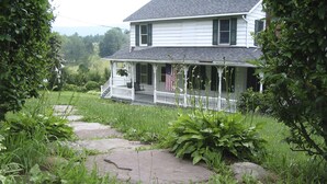 A two-story house with a porch, surrounded by lush greenery and a stone pathway.