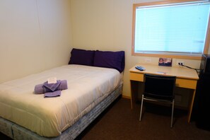 A single bed with a white comforter and purple pillows, a desk with a chair, and a window with blinds. - Target Hospitality-Pecos North Lodge (Pecos)