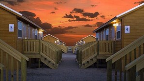 Une rangée de cabanes en bois avec des portes numérotées, un chemin de gravier et un ciel au coucher du soleil.