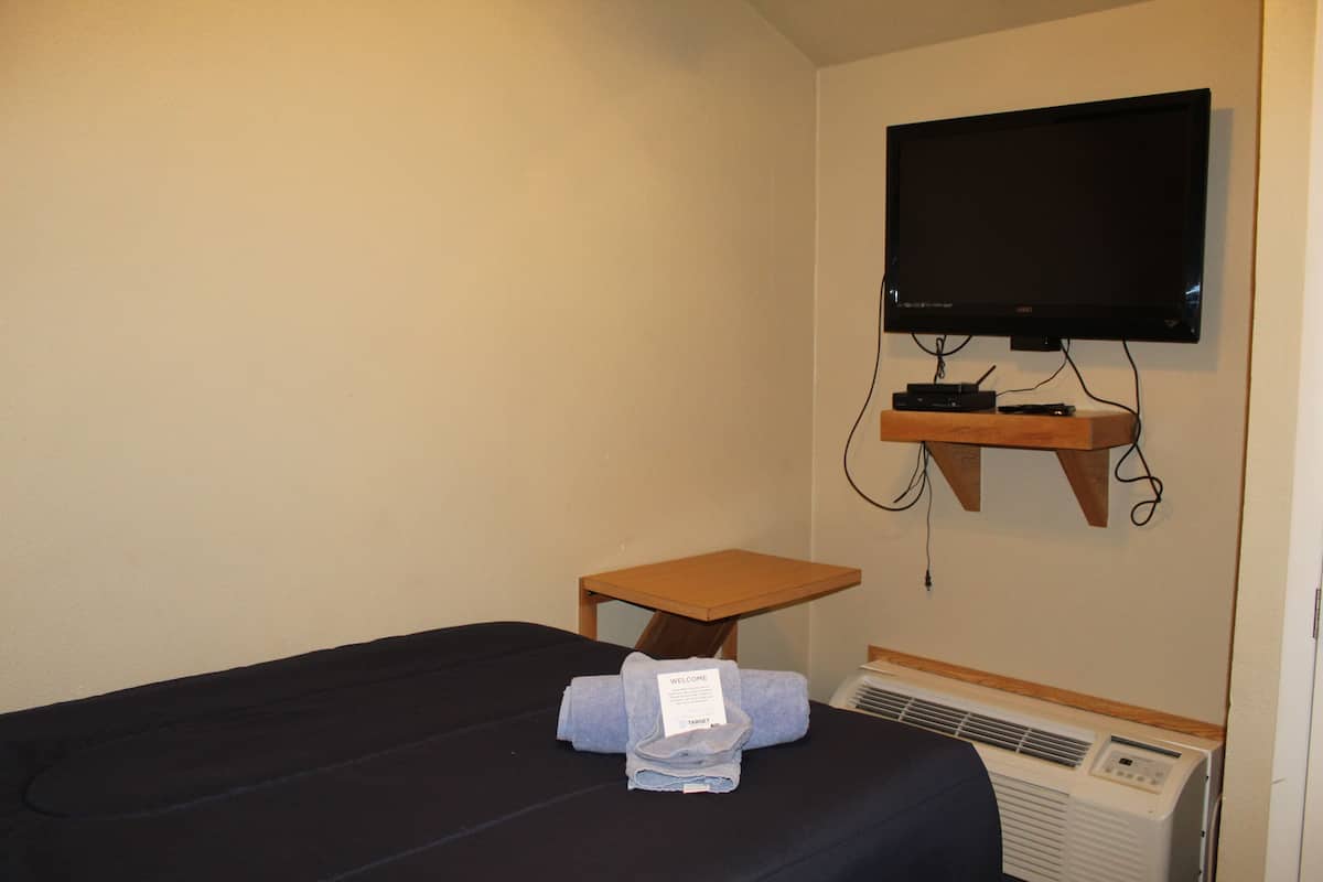 A hotel room with a single bed, a small wooden table, a television mounted on the wall, and a wall-mounted air conditioner.