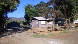 A small, single-story house with a sloped roof, surrounded by a wooden fence and a metal gate.