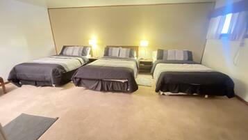 Habitación de hotel con tres camas individuales, cada una con una mesita de noche y lámpara. El suelo está alfombrado y hay una pequeña alfombra frente a las camas.