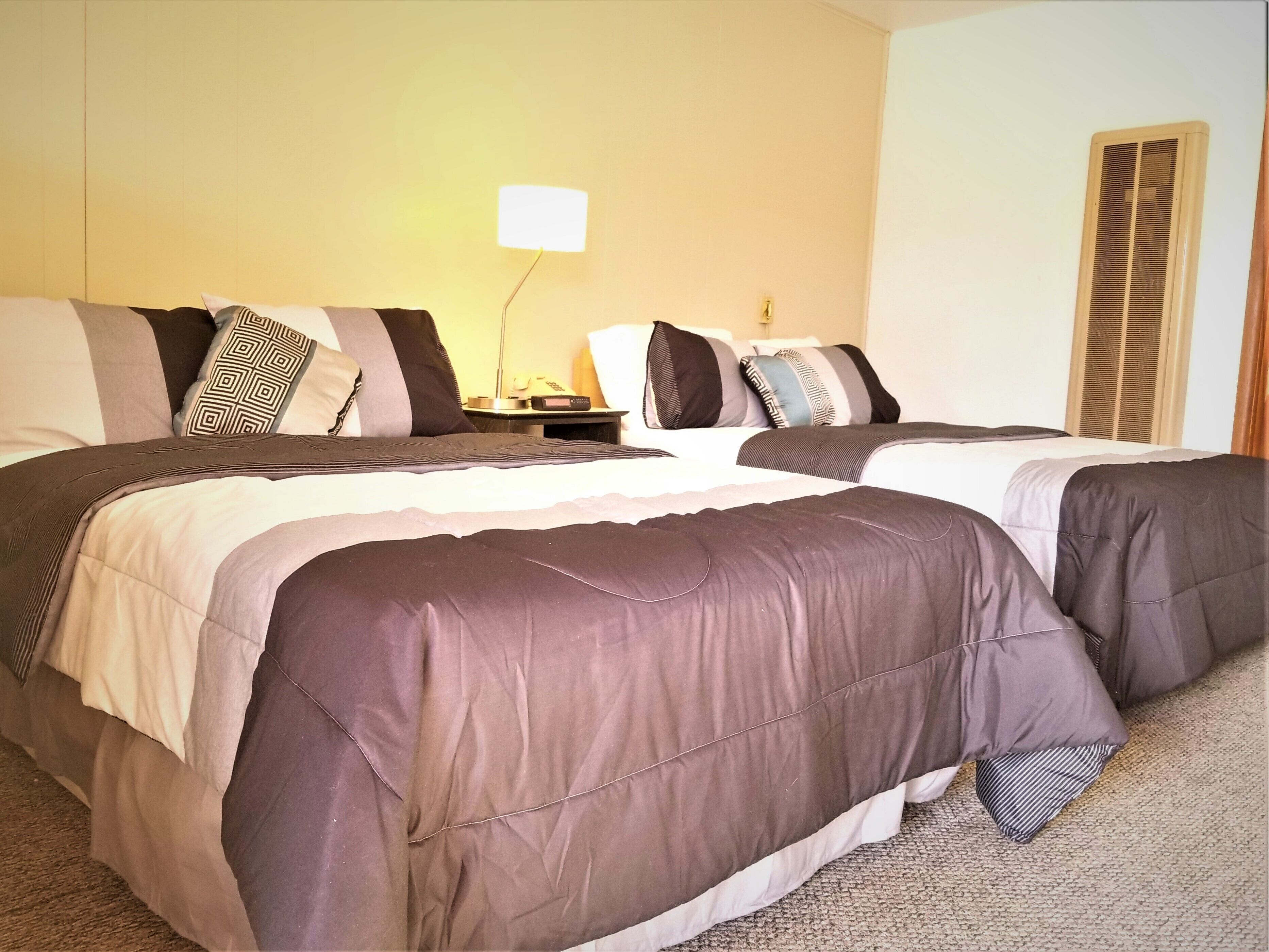 Habitación de hotel con dos camas, cada una con una colcha oscura y varios almohadones. Hay una mesita de noche con una lámpara y un control remoto.