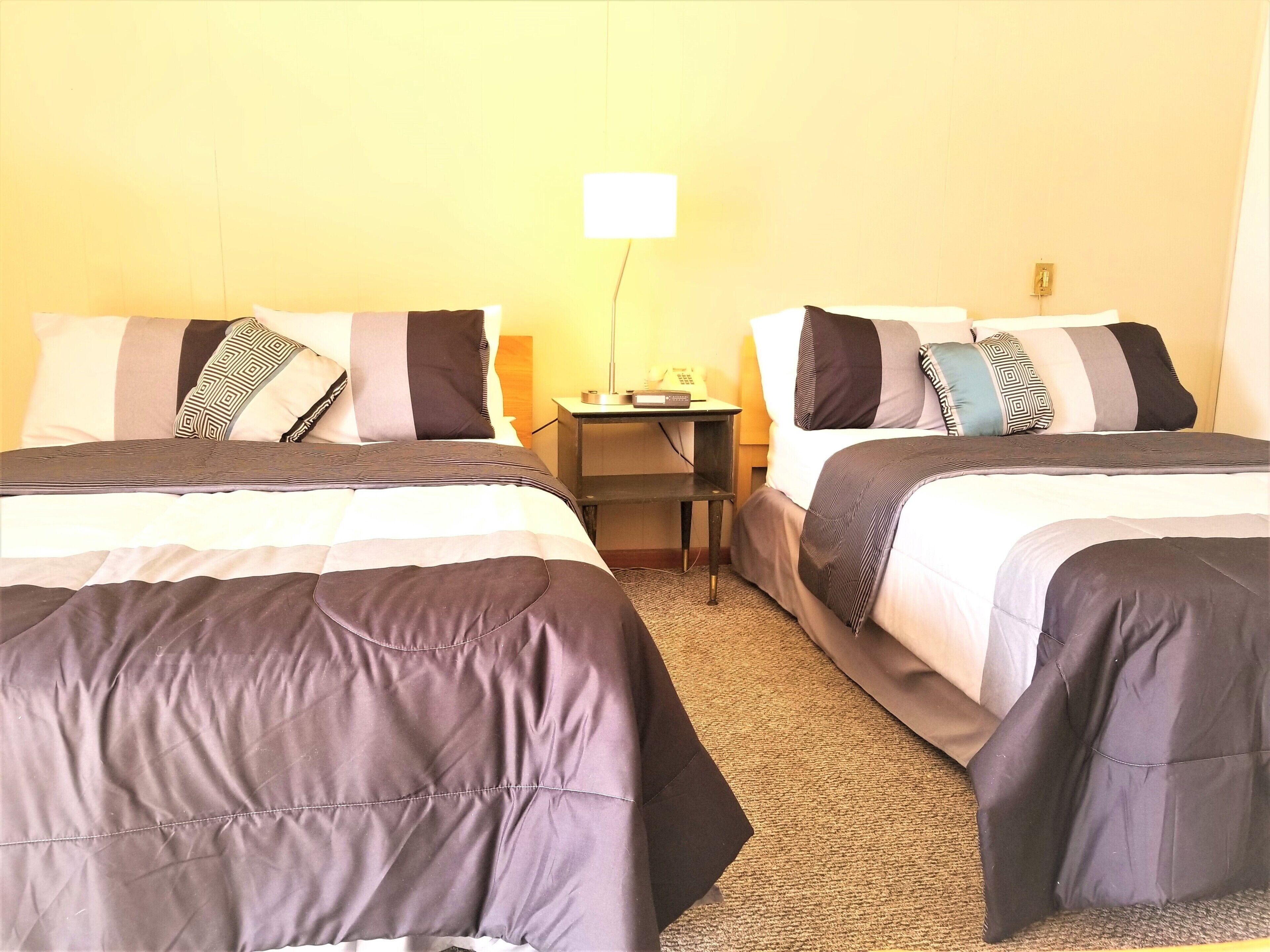 Habitación de hotel con dos camas, cada una con una colcha a rayas grises y negras, una mesita de noche con lámpara y un teléfono en la pared.
