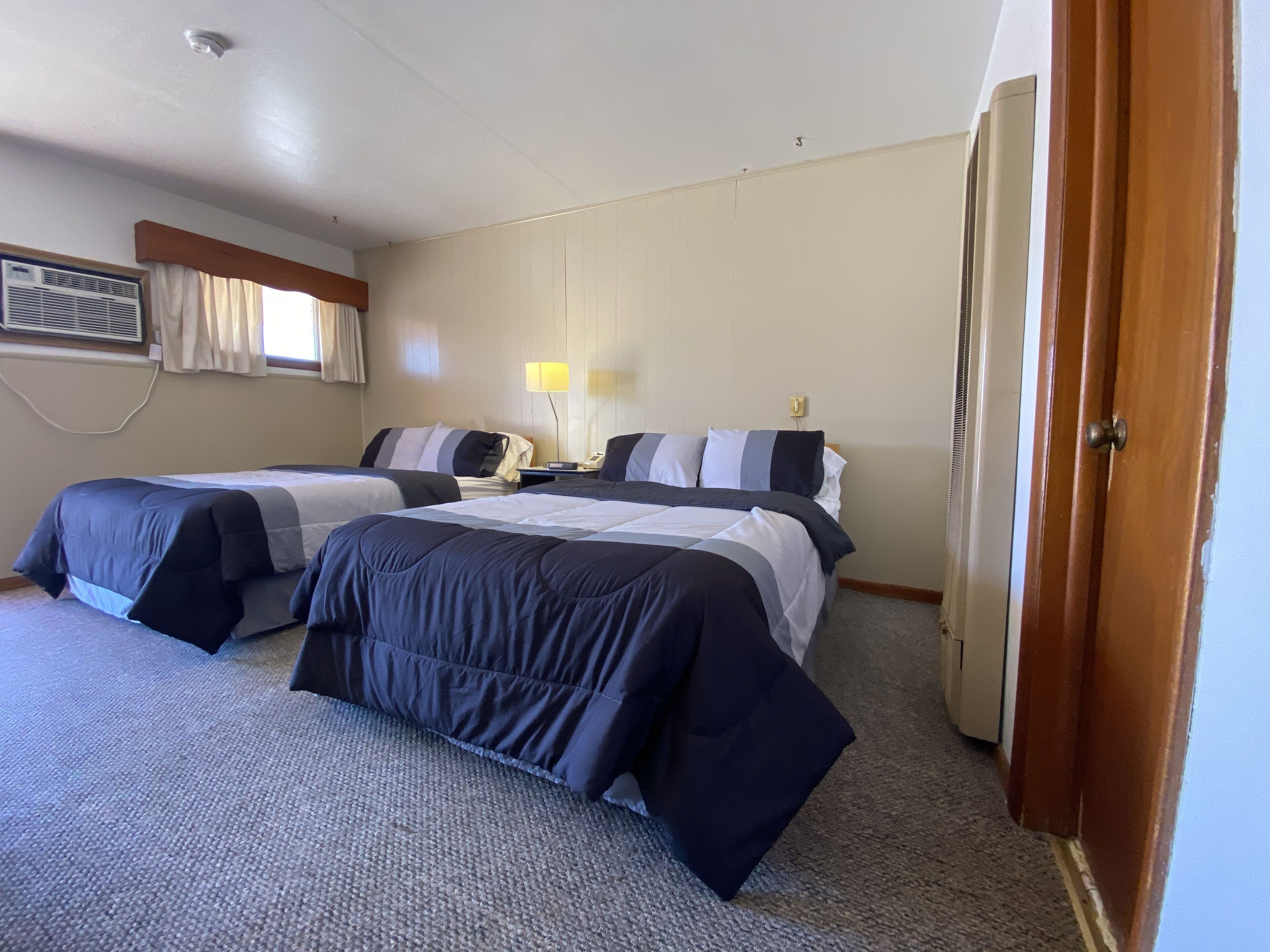 A hotel room with two beds, a desk, a lamp, and an air conditioning unit.