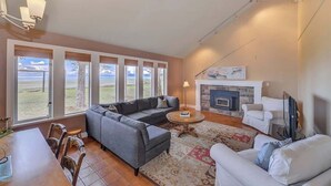 A spacious living room with a gray sectional sofa, a round coffee table, and a fireplace. Large windows offer a view of the outdoors. - Kye Bay Winter Beach House (Comox)