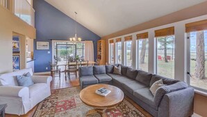 A spacious living room with a large gray sectional sofa, a white armchair, and a wooden coffee table. A patterned rug covers the floor, and the room has large windows offering a view of the outdoors. - Kye Bay Winter Beach House (Comox)