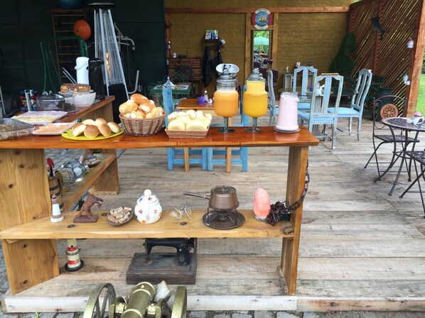 A rustic outdoor dining area with wooden tables and chairs, various food items, and a vintage sewing machine. - Gato de Botas Flats & Suites (Nova Friburgo)