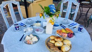 Un tavolo rotondo con tovaglia di pizzo bianco, un vaso con fiori e diversi cibi tra cui pane e frutta.