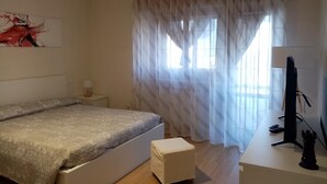 Una camera da letto con un letto, una scrivania, una sedia, una lampada, un televisore e una finestra con tende leggere.