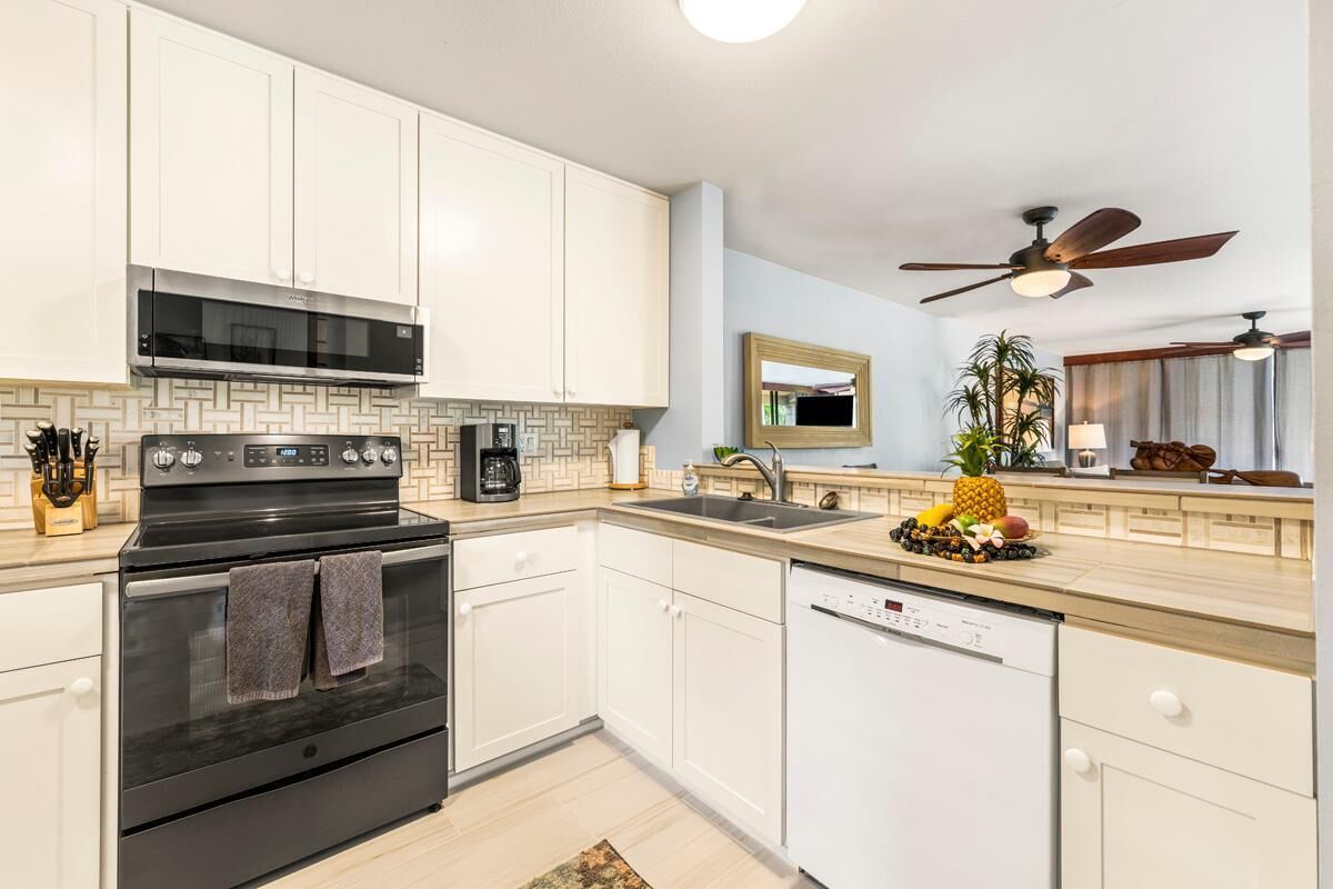 Une cuisine moderne avec des armoires blanches, une cuisinière noire et un lave-vaisselle. On y trouve également un ventilateur de plafond, un four à micro-ondes et un évier sur lequel est posé un panier de fruits.