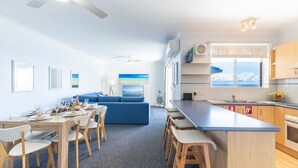 A modern kitchen with a central island, a dining area with a table and chairs, a blue sofa, and a large window with a view of the sea. - Kiah 12, 53 Victoria Pde - Panoramic water views, Wifi and Air Con (Nelson Bay)