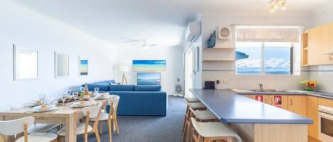 Une cuisine moderne avec un îlot central, un coin repas comprenant une table et des chaises, un canapé bleu et une grande fenêtre donnant sur la mer.