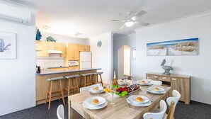 A modern kitchen with wooden cabinets, a microwave, and a refrigerator. There is a dining area with a wooden table set for four, surrounded by white chairs. A ceiling fan is visible above the table. - Kiah 12, 53 Victoria Pde - Panoramic water views, Wifi and Air Con (Nelson Bay)